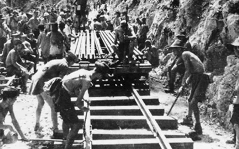 Black-and-white wartime photograph of labourers laying railway track by hand through a rock cutting.