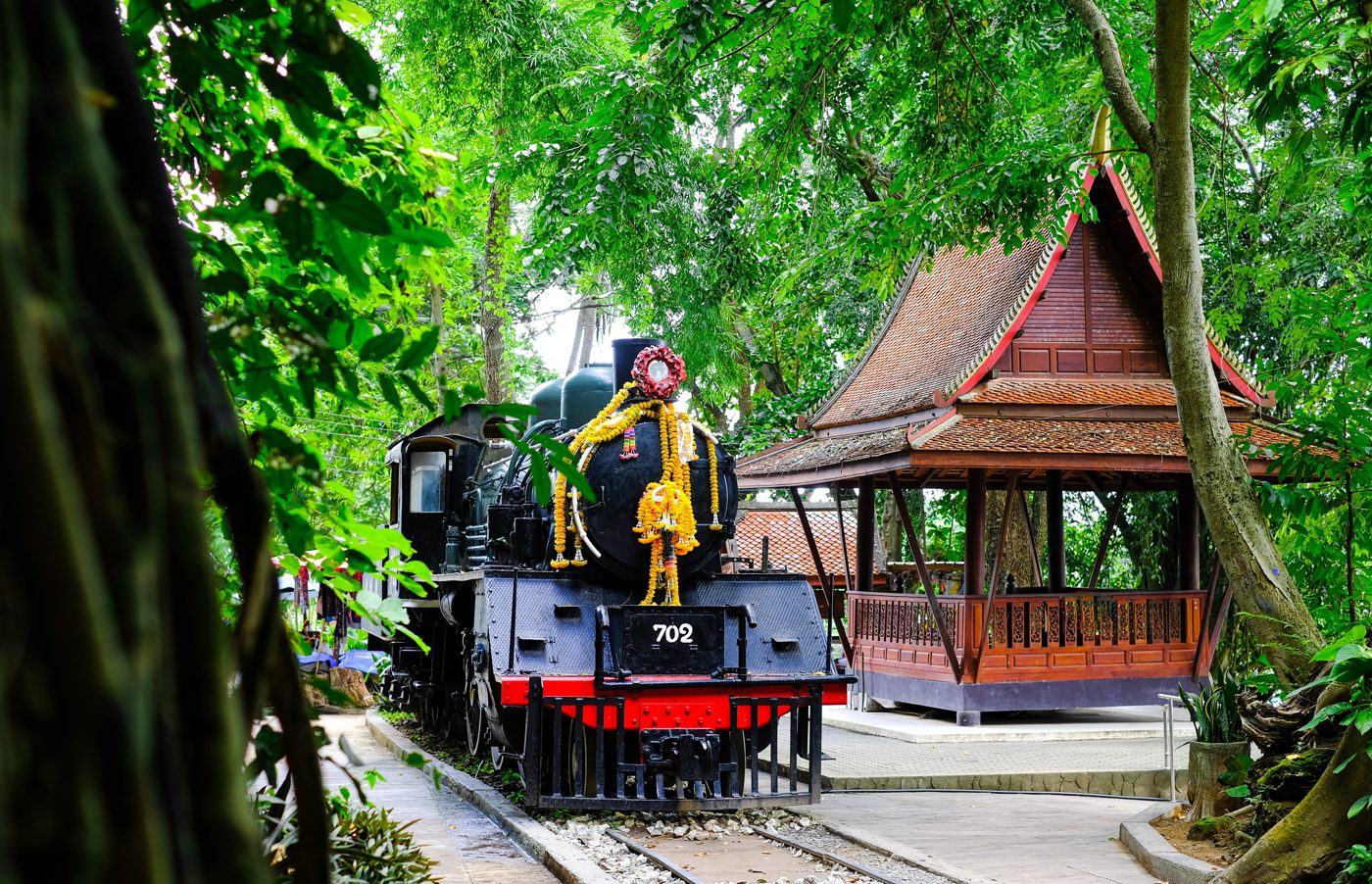 A preserved black steam locomotive draped with a marigold garland, displayed as a memorial beside a Thai pavilion in the jungle.